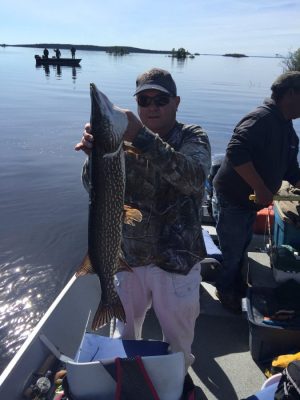 A man on a boat holds a large fish vertically, with another boat visible in the background on a calm body of water.