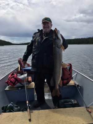 Person on a boat holding a large fish with a fishing rod nearby, against a background of water and trees.