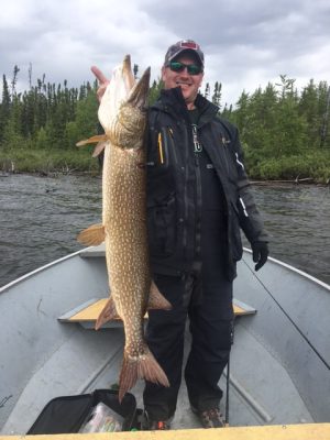 Person on a boat holding a large fish vertically with one hand, wearing dark outdoor clothing and sunglasses, trees in the background.