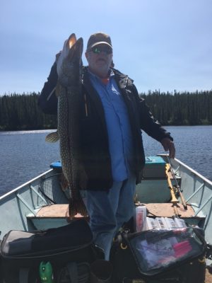 Man on a boat in a lake holding a large fish vertically, with fishing gear and trees in the background.