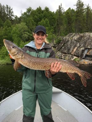 Person in a green outfit and black cap holding a large fish on a boat in a wooded area.