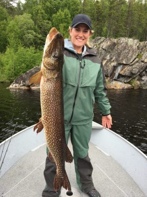 Person on a boat holding a large fish, standing in front of a rocky, forested shoreline.