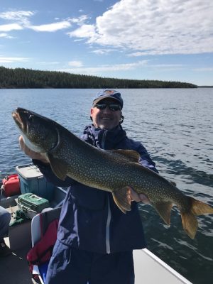 Person holding a large fish on a boat, with a lake and forested shoreline in the background under a partly cloudy sky.