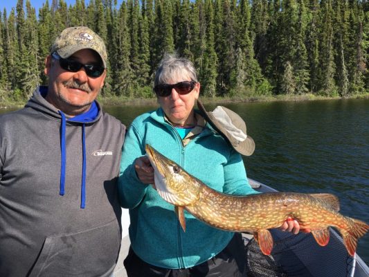 Two people on a boat hold a large fish together, with a forested shoreline in the background.