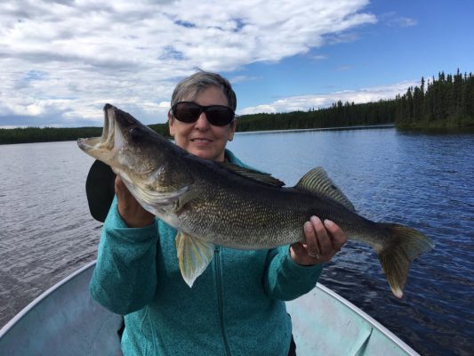 Person in a boat holding a large fish, smiling, with a forested shoreline and cloudy sky in the background.