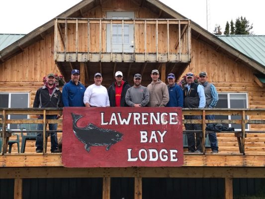 A group of nine men standing on a wooden lodge deck with a sign reading "Lawrence Bay Lodge" featuring a fish graphic.