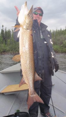Person holding a large fish on a boat, surrounded by water and trees under a cloudy sky.