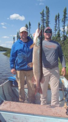 Two men in a boat hold a large fish upright. Trees and a lake are visible in the background under a blue sky.