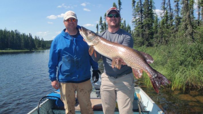 Two men in a boat on a lake, one holding a large fish. Forested shoreline in the background under a clear sky.