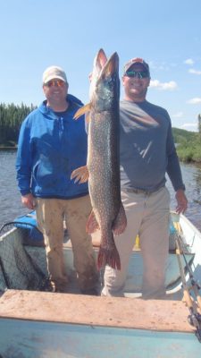 Two men on a boat holding a large fish vertically. Fishing rods and net are visible. Trees and sky form the background.