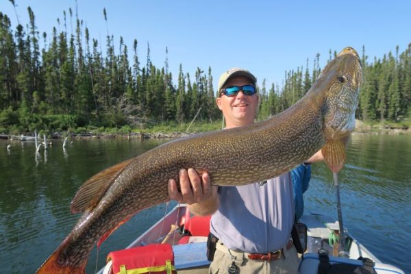 A person on a boat holds a large fish with both hands; trees and water are visible in the background.
