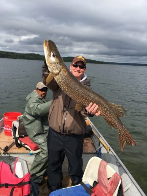 Two men in a small boat on a lake; one holds a large fish with both hands, smiling at the camera.