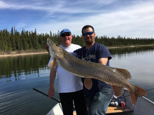 Two men on a boat hold a large fish over the water with trees and a blue sky in the background.