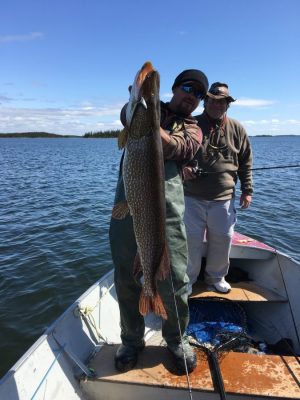 Two people on a small boat; one holds a large fish while the other stands behind, both on a lake under a clear sky.
