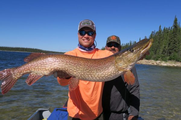 Man in sunglasses and a cap holds a large fish by the shore, with another man and trees in the background.