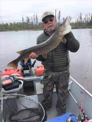 Man in a boat holding a large fish, wearing sunglasses and a cap. Fishing rod and net are visible. Forest and lake in background.