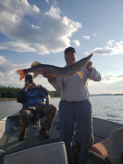 Two people on a boat; one holds a large fish while the other takes a photo. Trees and water are visible in the background.