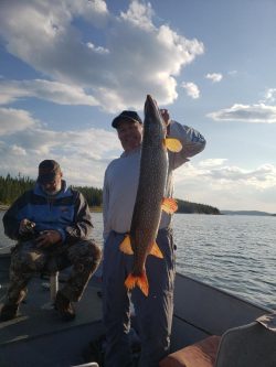 Person standing on a boat holding a large fish; another person is sitting and looking at something in their hands.