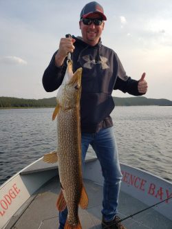 Man standing on a boat holds up a large fish with one hand and gives a thumbs up with the other.