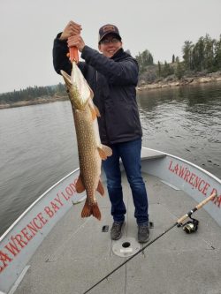 Person standing on a boat holding a large fish with both hands; fishing rod and water are visible in the background.