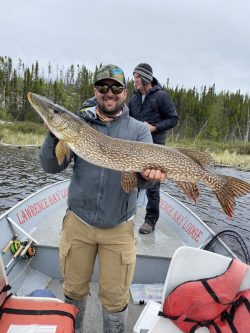 Man standing on a boat holding a large fish, with another person and trees in the background.