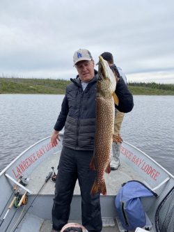 A man on a fishing boat holds up a large fish he caught, with fishing gear and another person in the background.