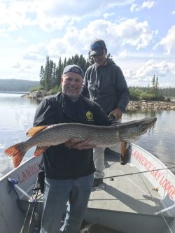 Two men stand on a boat; one holds a large fish with both hands on a lake surrounded by trees.