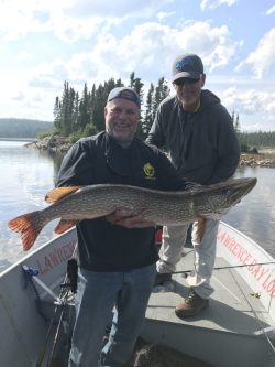Two men stand on a boat; one holds a large fish, with a lake and trees in the background under a partly cloudy sky.
