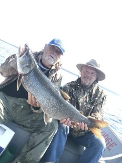 Two men sitting in a boat hold a large fish they caught, posing for a photo on the water.