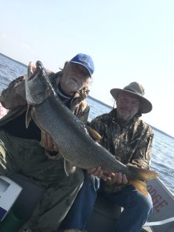 Two men sitting in a boat on a lake, holding a large fish together and looking at the camera.
