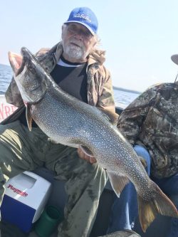 Older man in camouflage jacket and blue cap holding a large fish on a boat with another person beside him.