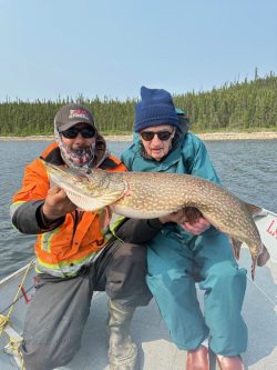 Two people in a boat hold a large northern pike fish over a lake with trees in the background.