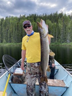 Man standing in a boat on a lake holding a large fish, with trees and cloudy sky in the background.