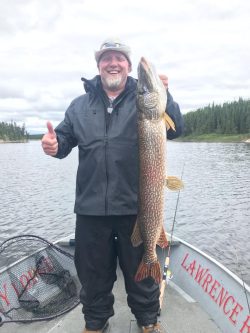 Man on a boat holding a large fish vertically with one hand and giving a thumbs up with the other, lake and trees in background.