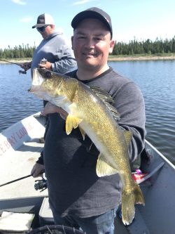 Man holding a large yellow fish on a boat with another person fishing in the background on a lake.