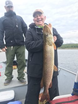 Smiling person on a boat holds a large fish upright, with another person standing behind them.