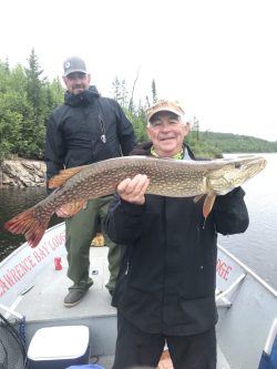Two men on a boat; one is smiling and holding a large fish, with trees and water in the background.