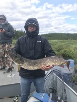Person in a black jacket holding a large fish on a boat, with another person standing in the background.