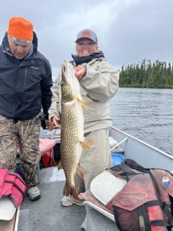 Person in rain gear holds a large fish on a boat, with another person and lake with trees in the background.