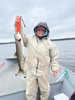 Person in rain gear holding a large fish on a boat in a lake under an overcast sky.
