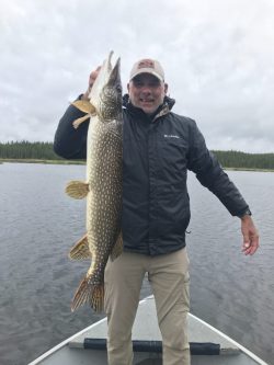 Man standing on a boat holds up a large Northern pike fish with both hands on a cloudy day.
