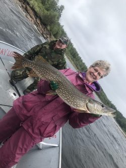 Smiling woman in a pink jacket holds a large fish on a boat, with a man in camouflage standing behind her.