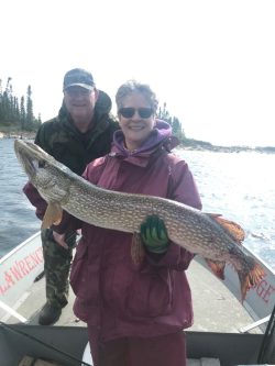 Two people on a boat, one holding a large fish, both smiling at the camera near a wooded shoreline.