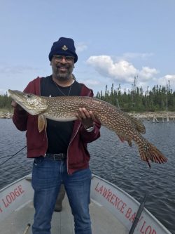 Man standing on a boat holding a large fish with both hands, smiling, with water and trees in the background.