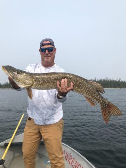 Man standing on a boat holds a large fish with both hands over a lake, with trees visible in the background.