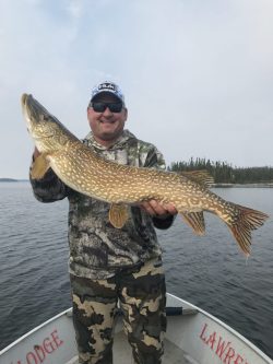 Man standing on a boat holds a large fish with both hands; lake and trees visible in the background.