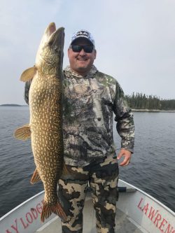 Man standing on a boat holding a large fish upright, with water and trees visible in the background.