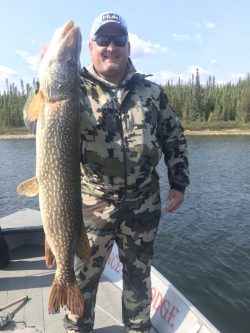Man in camouflage clothing on a boat holds up a large fish with a lake and trees in the background.