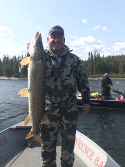 Person in camouflage clothing holding a large fish on a boat, with another person and trees visible in the background.