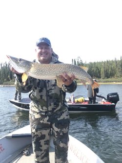A person in camouflage clothing holds a large fish on a boat; another boat and trees are visible in the background.